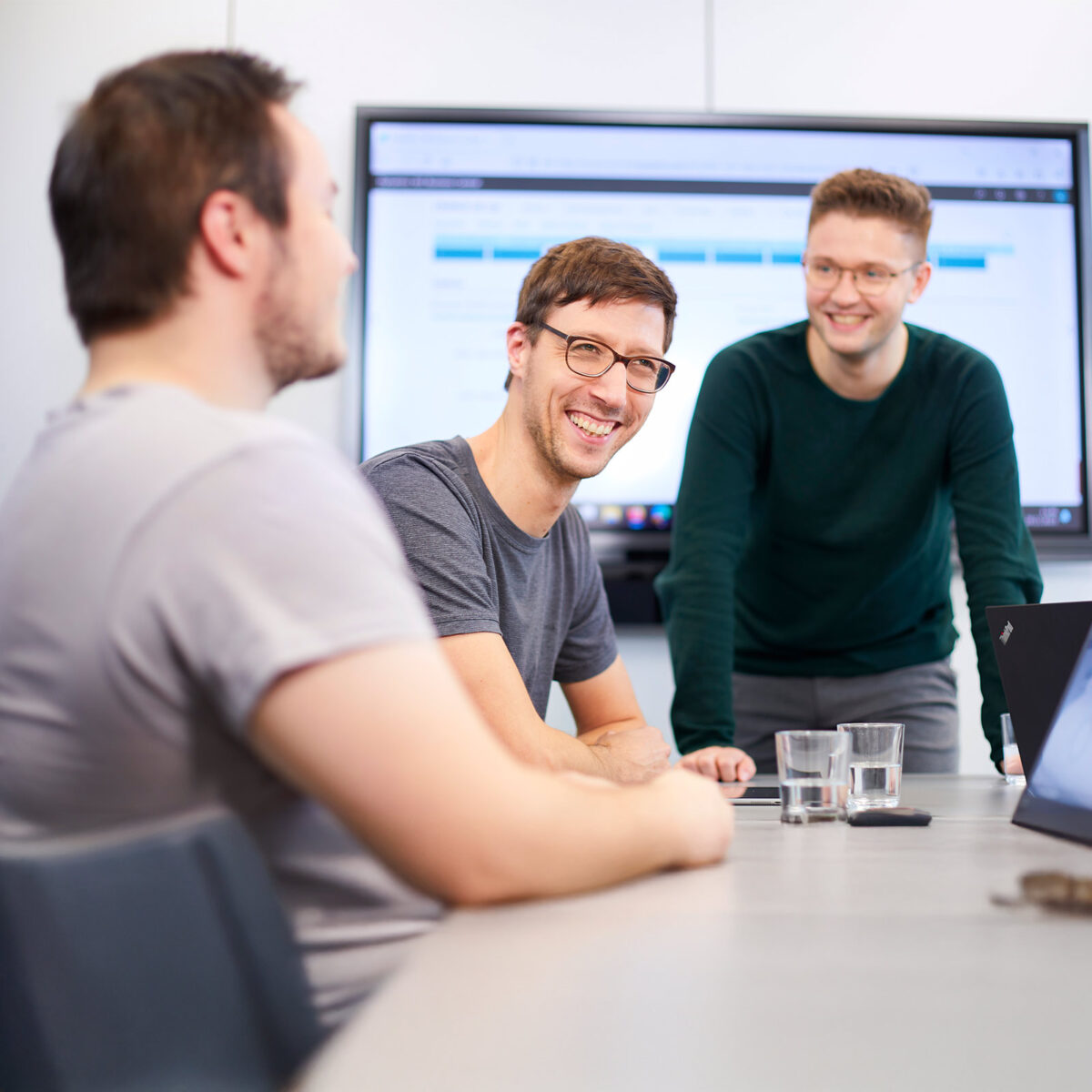 Zwei Männer sitzen an einem Tisch, ein Mann steht davor. Sie lachen. Im Hintergrund steht ein Monitor, auf dem Business Central zu sehen ist.